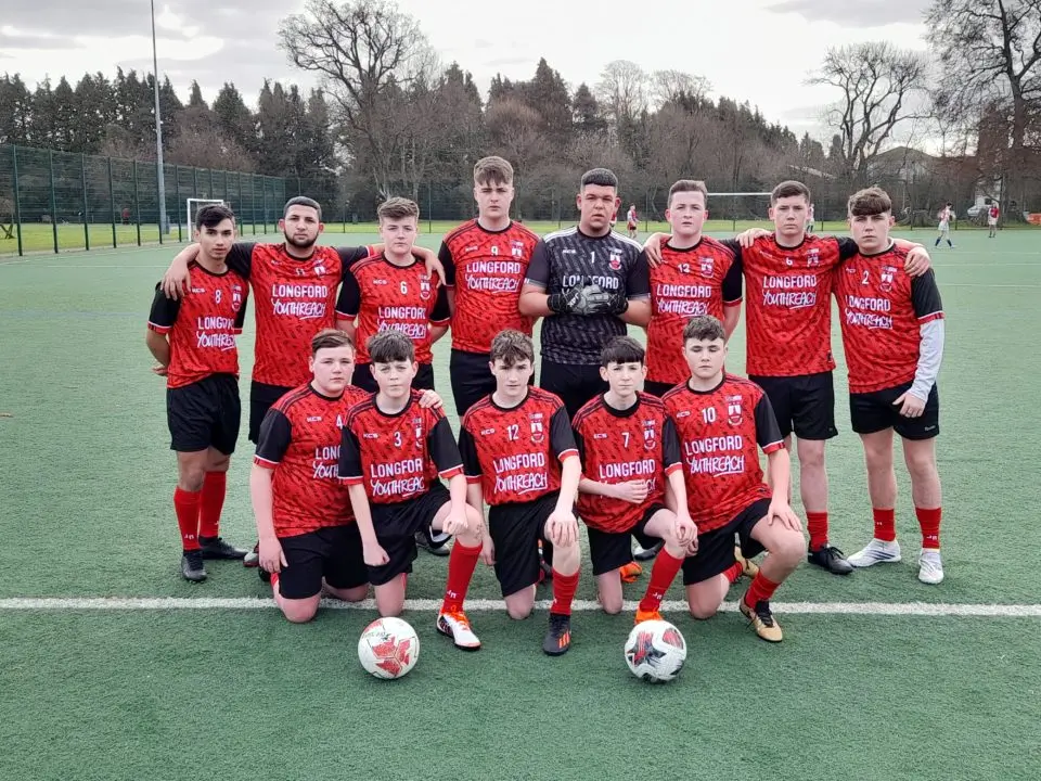 13 boys wearing red football kit. 5 boys are kneeling at the front of the picture and 8 are standing behind.