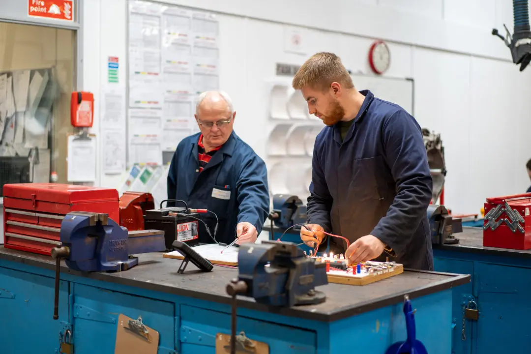 An electrical apprentice and their tutor standing at a workbench testing equipment, in an electrical workshop. They are standing at blue workbench with two table top vices in the foreground of the image.