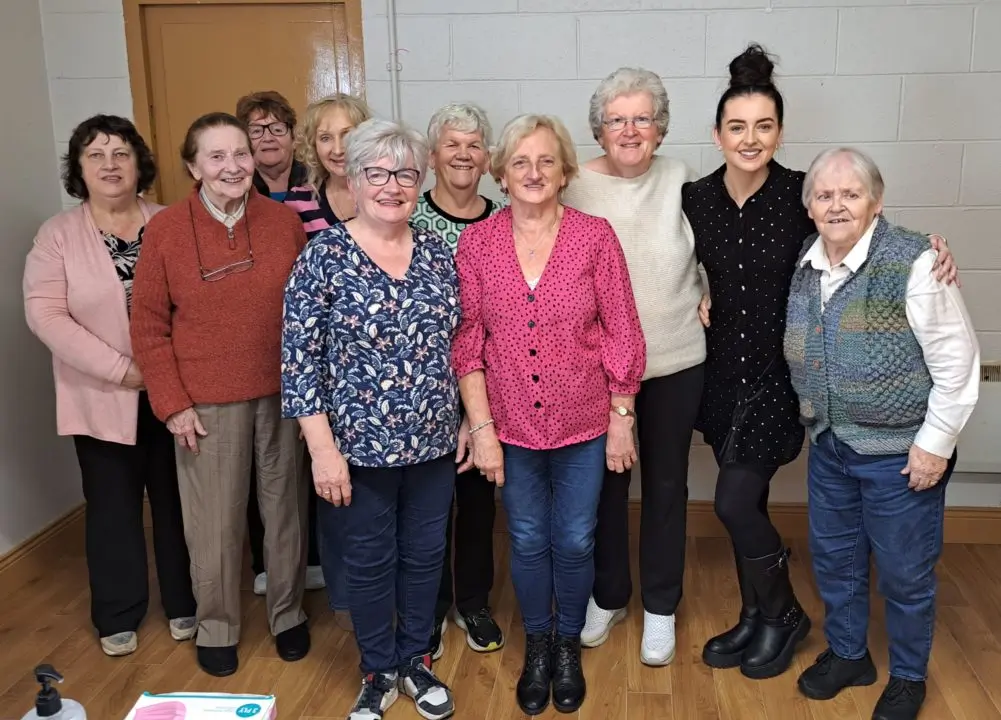 Nine older learners standing in front of a white painted brick wall, smiling and facing the camera. The tutor is standing at the end with her arm around one of the learners.