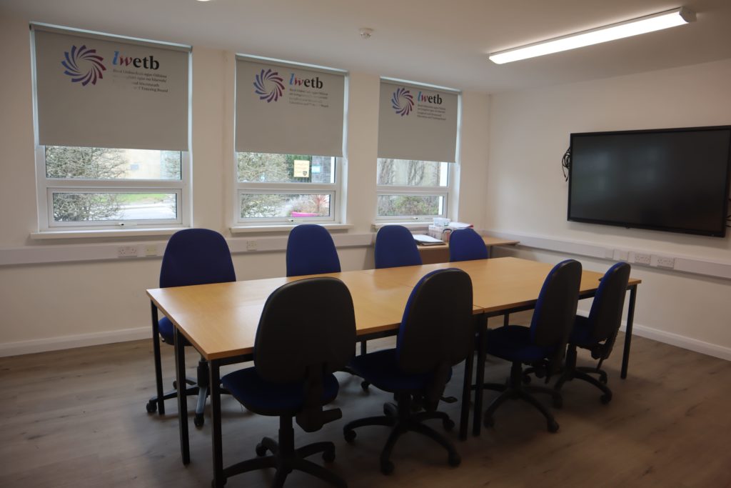 An empty classroom with a boardroom style rectangular table with four chairs on either side. Behind the table are three windows with the roller blinds half closed, the blinds show the LWETB logo on each.
