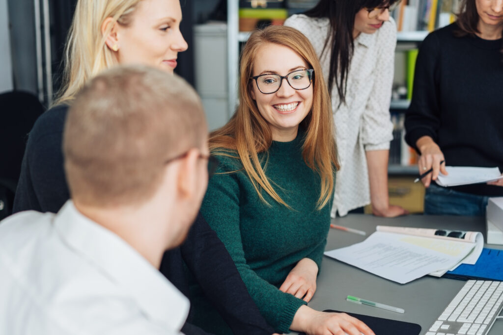 A young woman smiling and wearing glasses surrounded by people in an office.