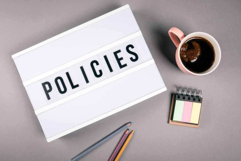 A lightbox displaying the word POLICIES sits on a grey surface next to a pink coffee cup, a small notepad with pastel sticky notes, several coloured pencils, and hints of quality assurance in the organised workspace.