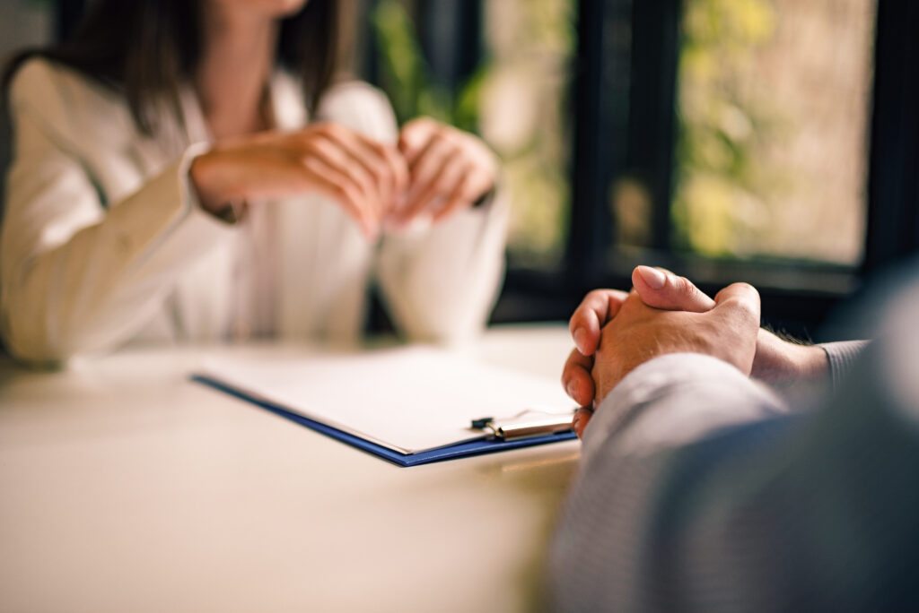 Two people sit opposite each other at a table, one with hands clasped, the other gesturing. A clipboard with papers lies on the table, suggesting a Quality Assurance meeting or interview setting.