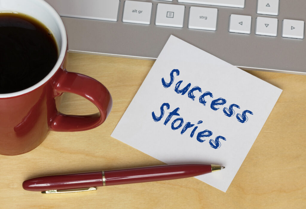 A red coffee mug, a red pen, and a note with Success Stories—highlighting Adult Literacy—sit on a wooden desk next to a silver keyboard.