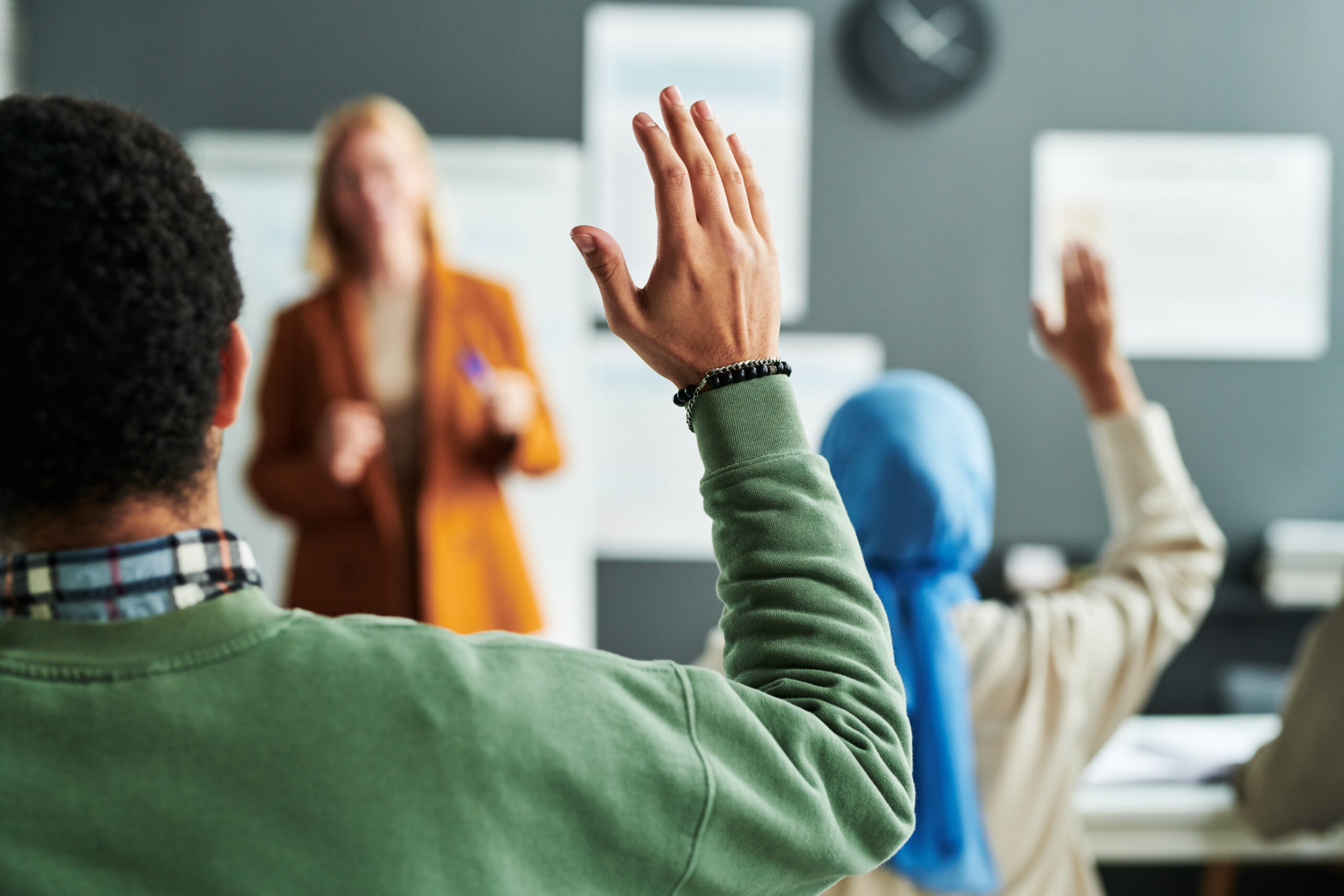 Part of back of young male student in green jumper with his hand raised a teacher standing at the top of the classroom.