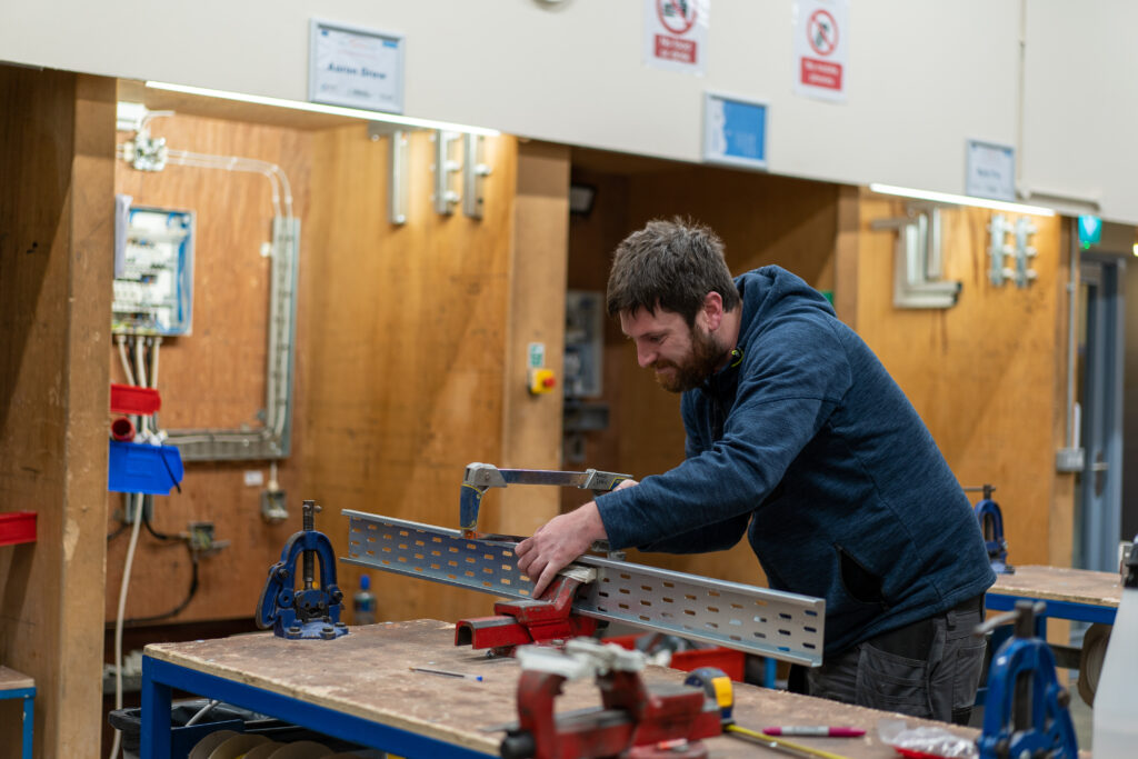 An apprentice is standing in front of a workstation in an Electrical workshop. He is focussing on marking a metal sheet with a red marker.