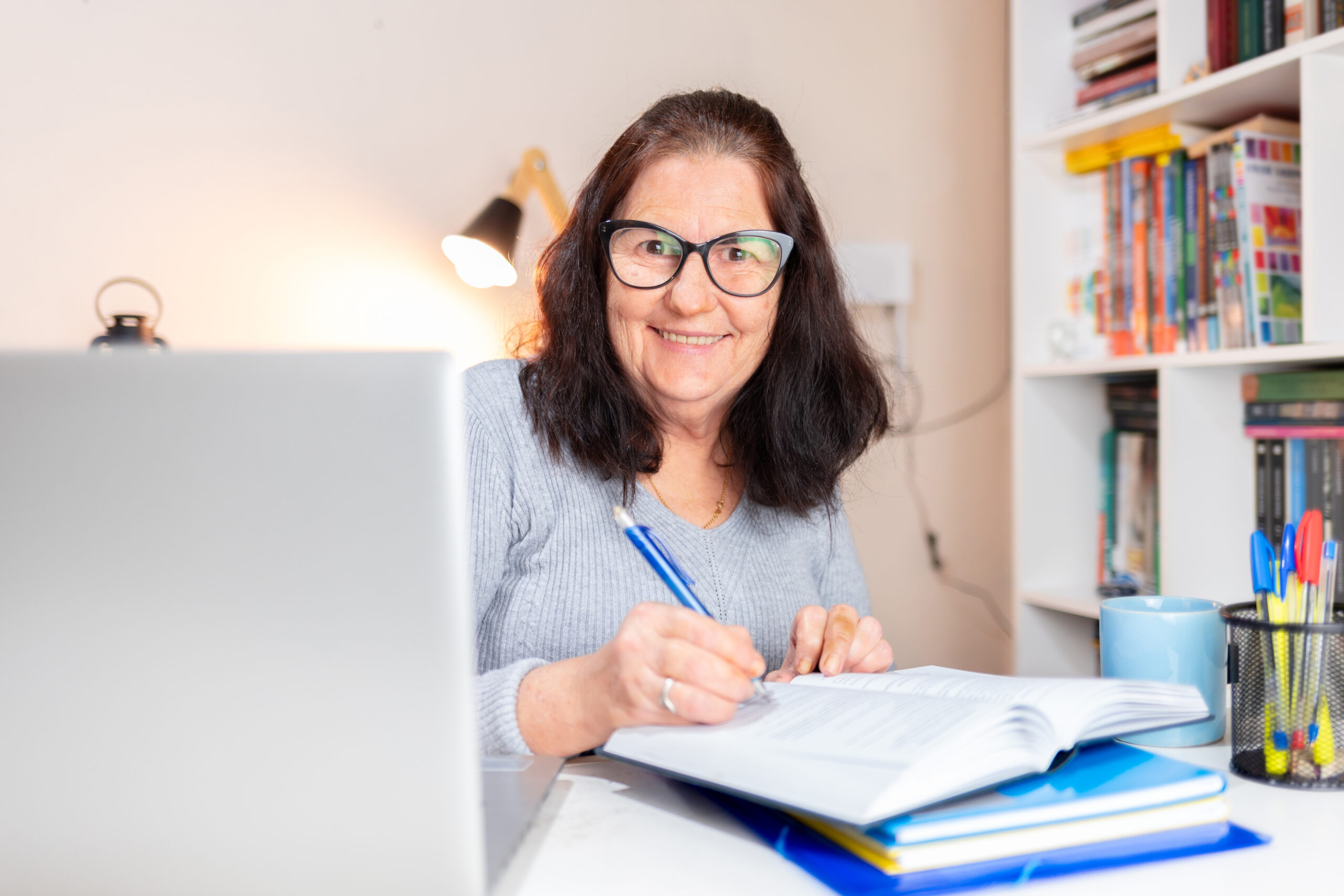 A woman sitting at a desk with an open laptop. She is writing in a diary, facing the camera and smiling.