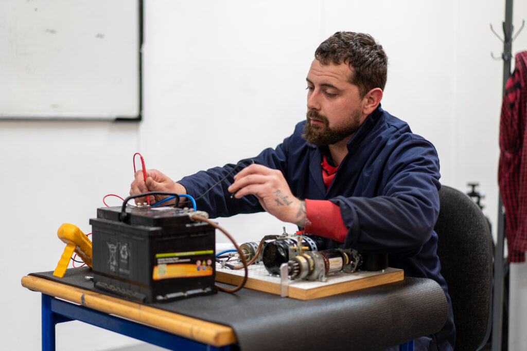 A motor apprentice is seated, inspecting the motor battery on the desk in front of him. He is concentrating on the battery and wearing blue overalls.