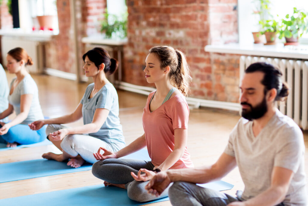A group of people sit cross-legged on yoga mats indoors, meditating with eyes closed and hands on their knees. The serene space, with brick walls and large windows, fosters mindfulness and Learner Support among participants.