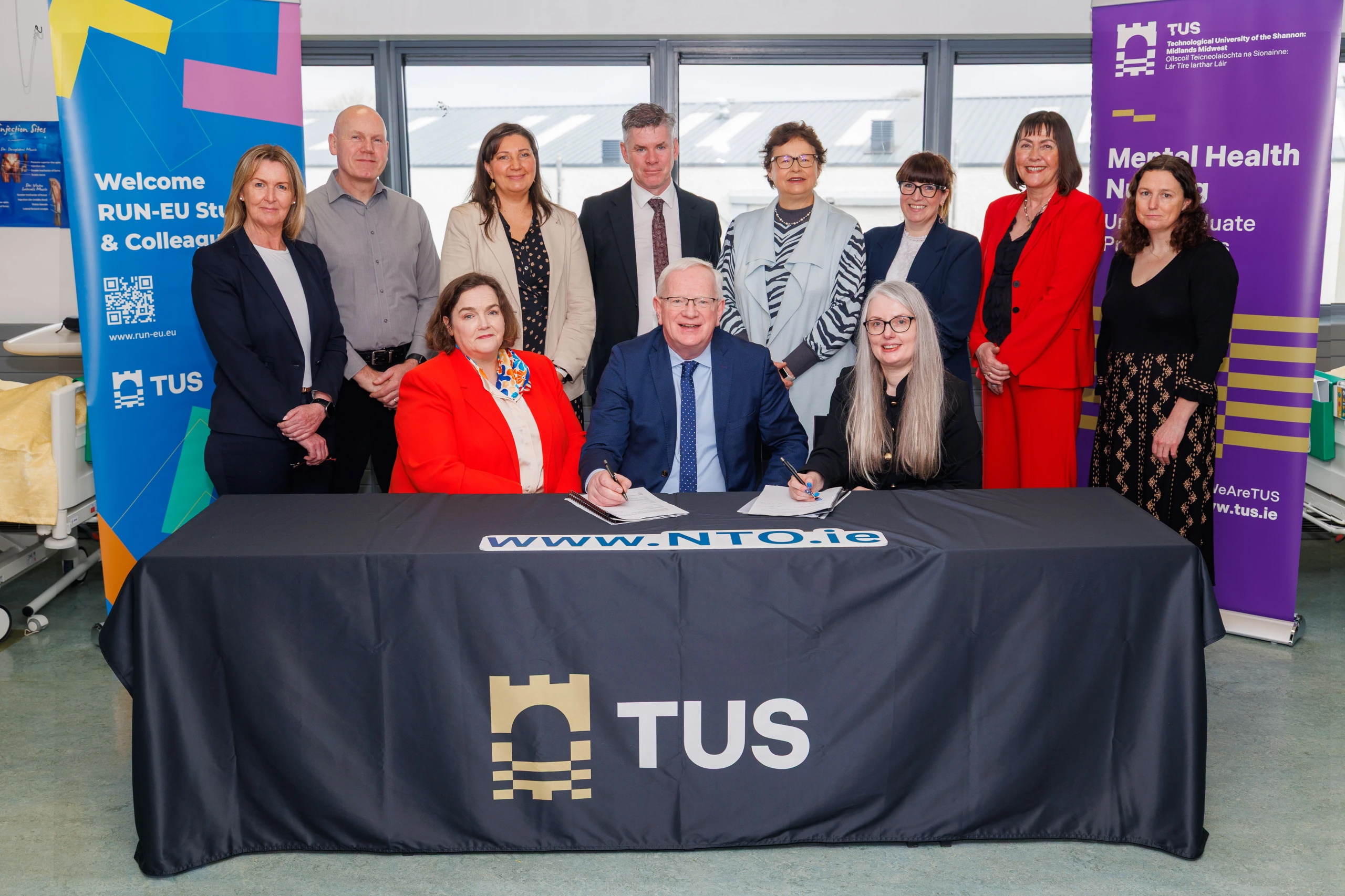 A group of eleven people pose indoors behind a table with a TUS-branded tablecloth; two people are seated, signing documents related to tertiary Nursing and Mental Health studies, whilst the rest stand behind them. LWETB Staff